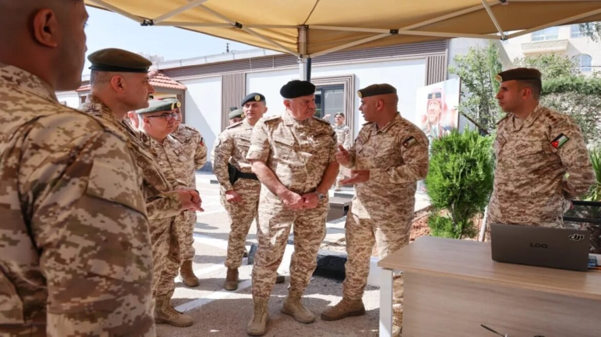 Group of soldiers in desert camouflage under a canopy, listening as a officer speaks near a laptop on a table outdoors.