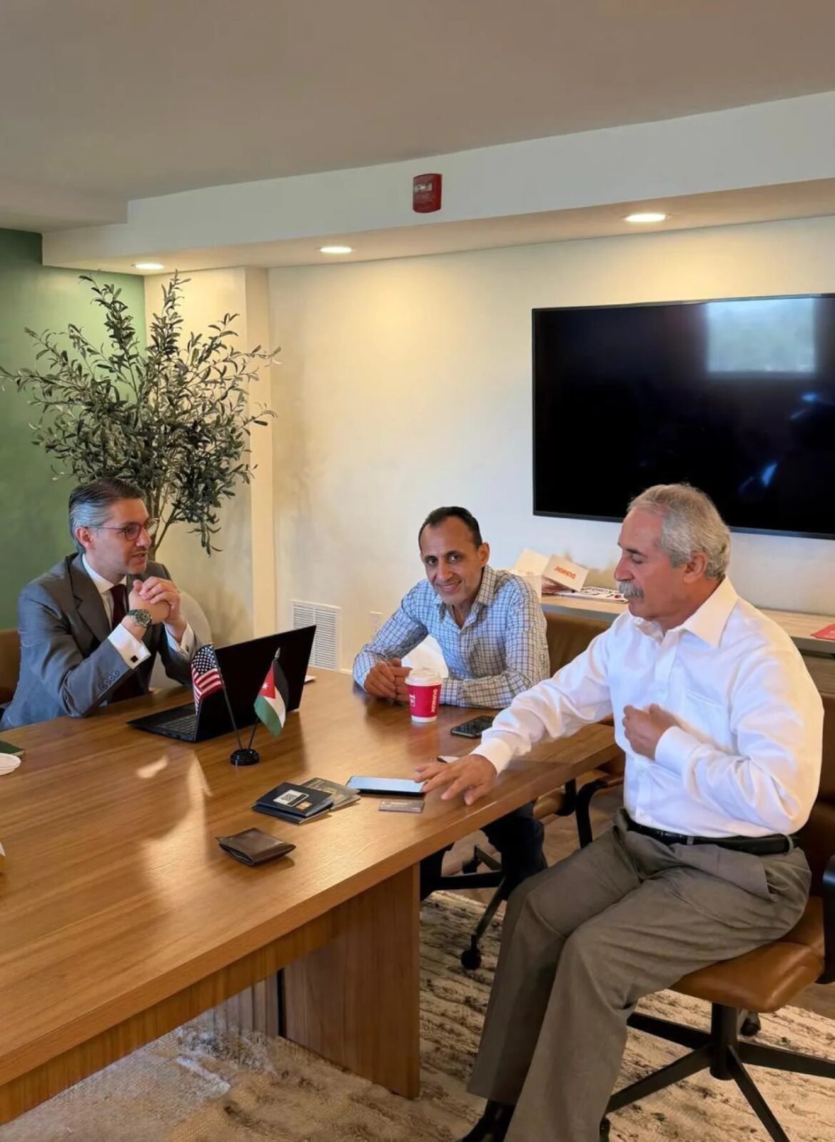 Three men in a conference room sit at a wooden table with laptops and small flags, discussing a meeting.