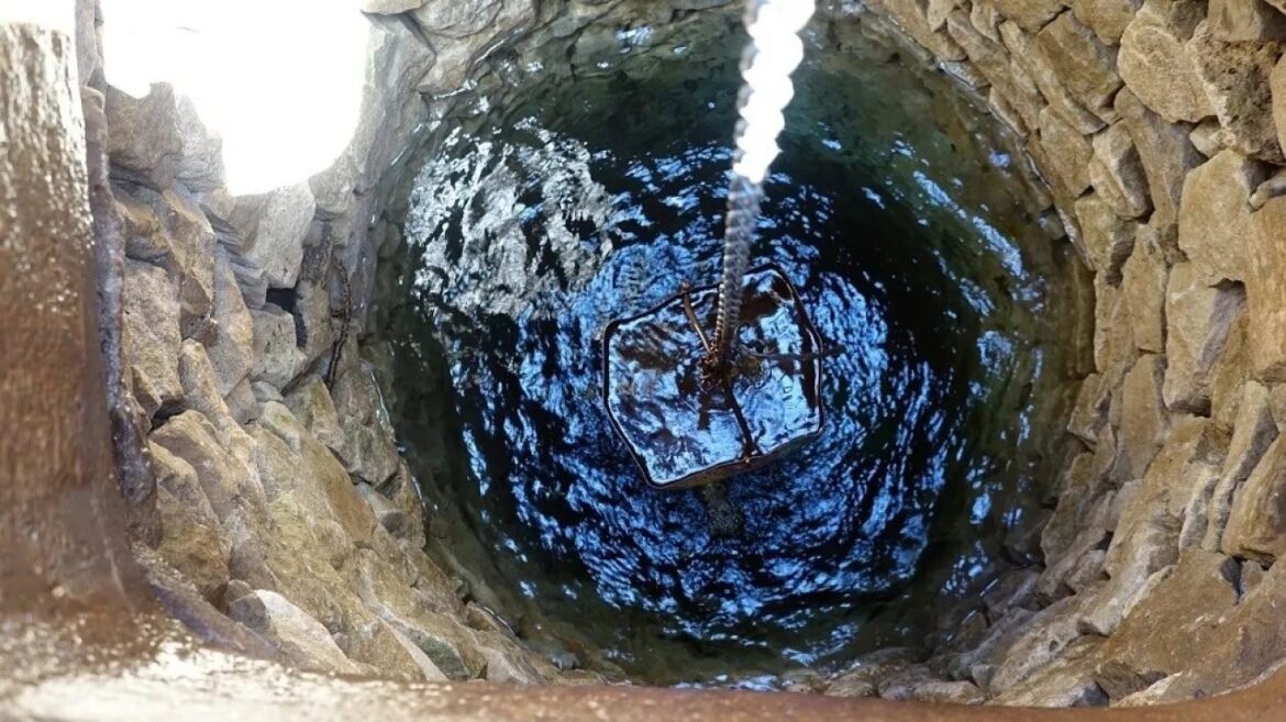 Rusty square metal cage hanging in a deep, stone-walled well with dark water at the bottom and a rope above it.