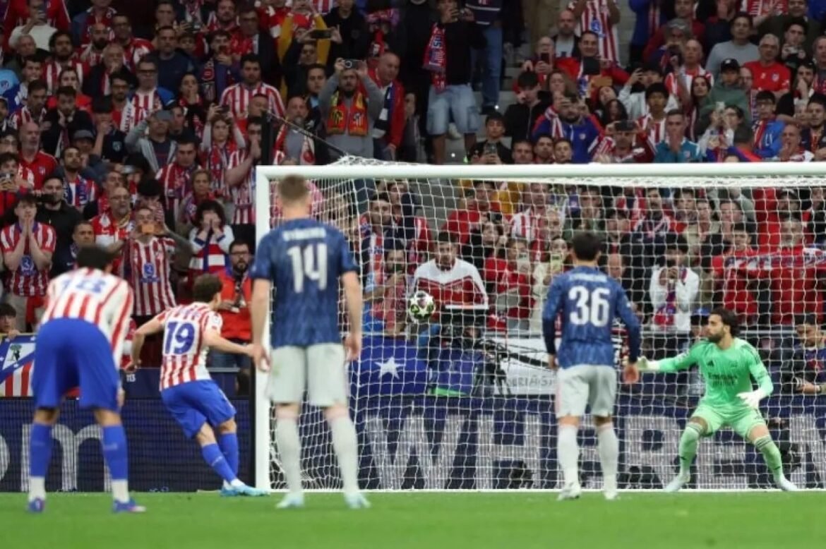 Soccer match with red-and-white striped players near the goal as the ball heads toward the net and a green-clad goalkeeper prepares to stop it.