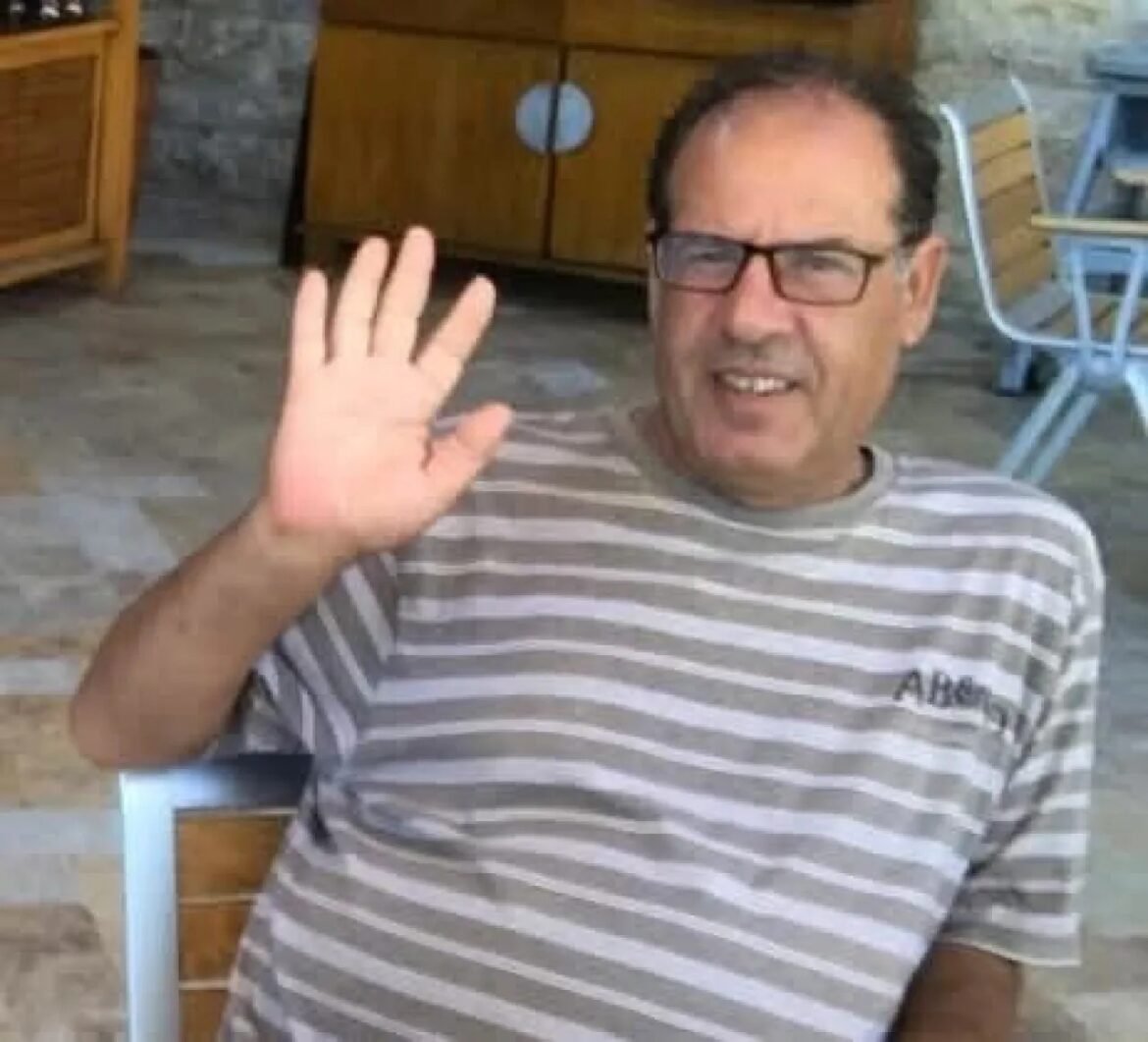 Middle-aged man wearing glasses in a striped shirt waves hello at the camera, seated on a patio with stone floor.