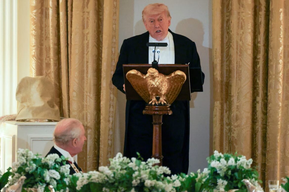 Donald Trump delivering a speech at a wooden podium shaped like an eagle, with gold curtains in the background and microphones in front of him.