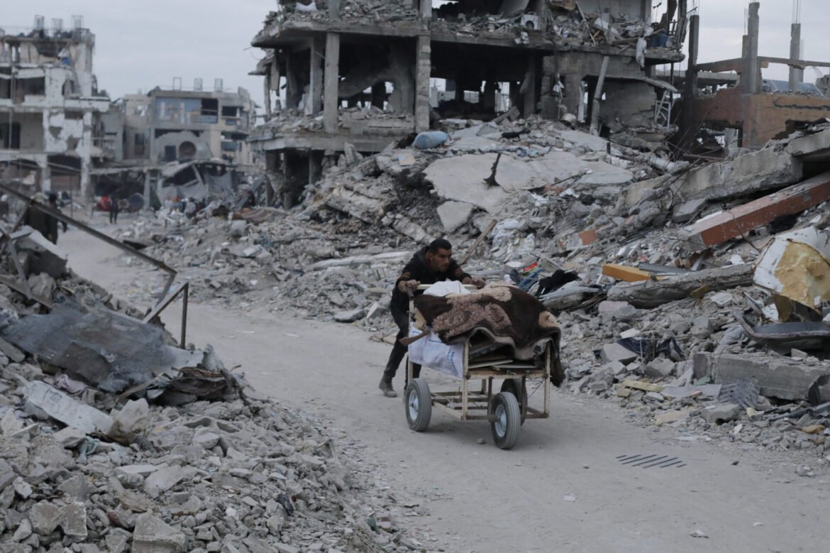 Man pushes a cart loaded with blankets through a street of rubble and destroyed buildings after a bombing or disaster.