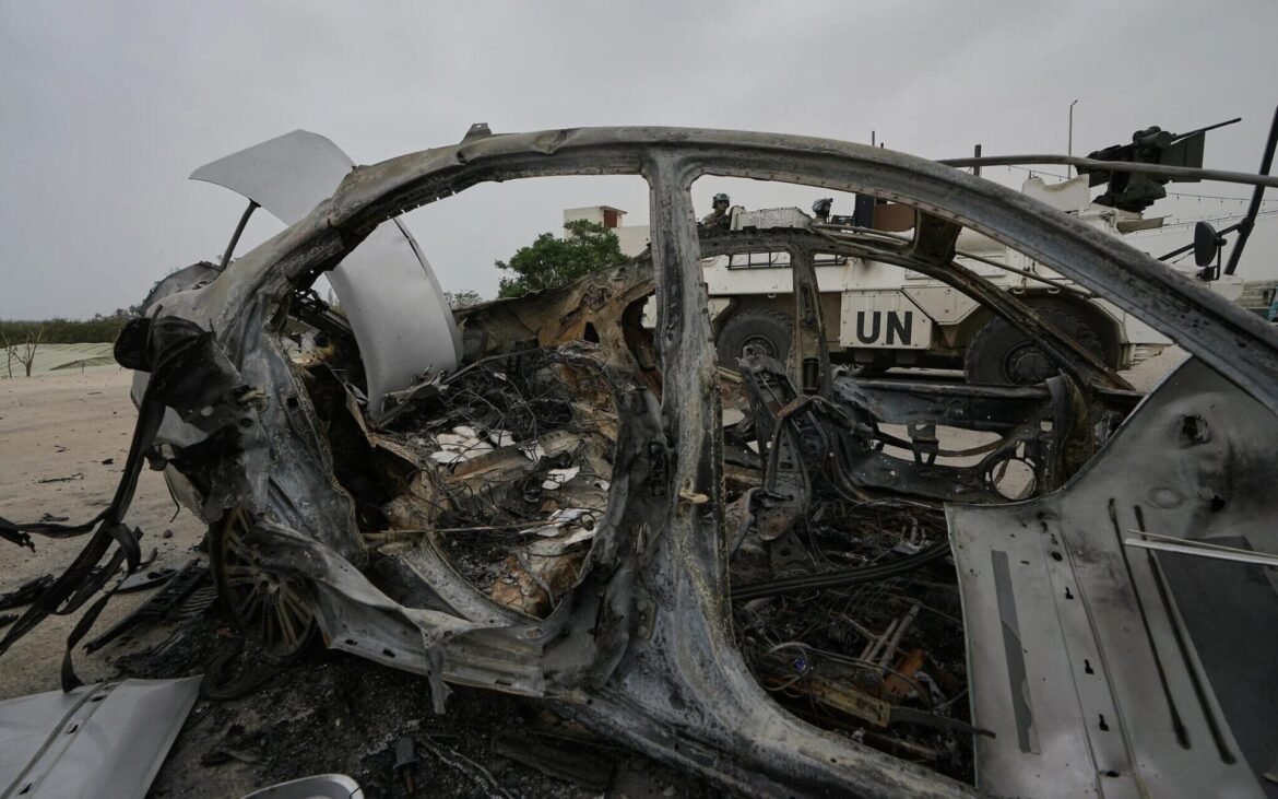 Charred, mangled car wreck in a desert area with a UN armored vehicle and soldiers visible in the background, indicating recent combat or explosion.