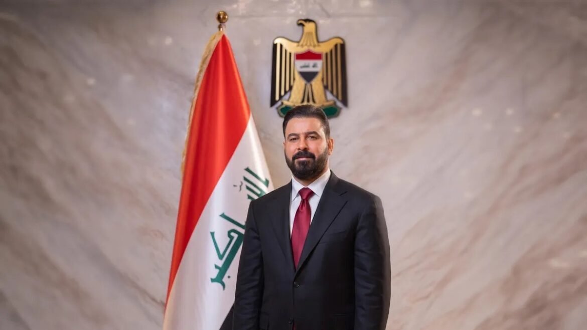Man in a dark suit and red tie stands in front of the Iraqi flag and national emblem in a formal setting.