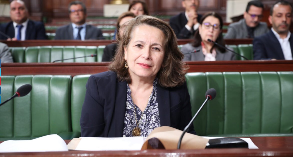 Woman in a navy blazer sits at a desk in a parliamentary chamber with a microphone and papers nearby.