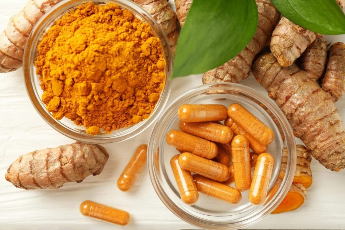 Top-down view of ground turmeric powder in a glass bowl with fresh ginger roots and orange turmeric capsules nearby, on a light wooden surface with green leaves for garnish.