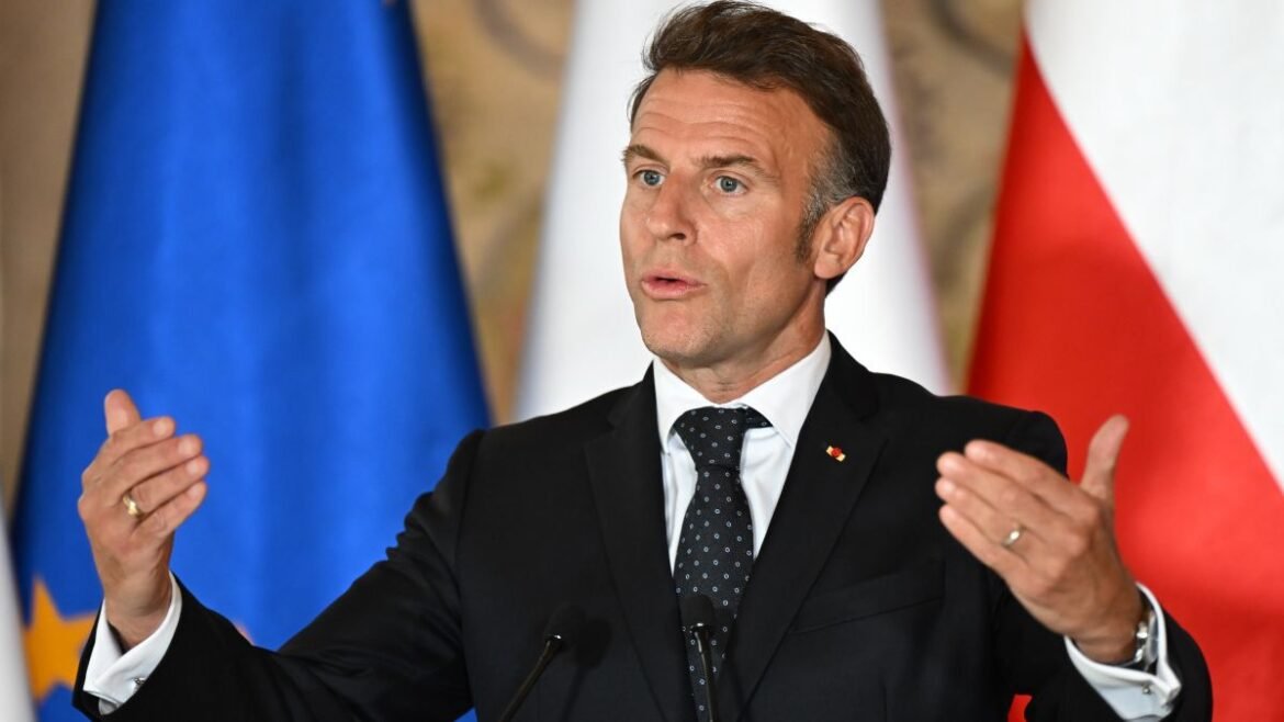 Politician in a dark suit and dotted tie speaks at a podium, gesturing with hands, with blue and red flags in the background.