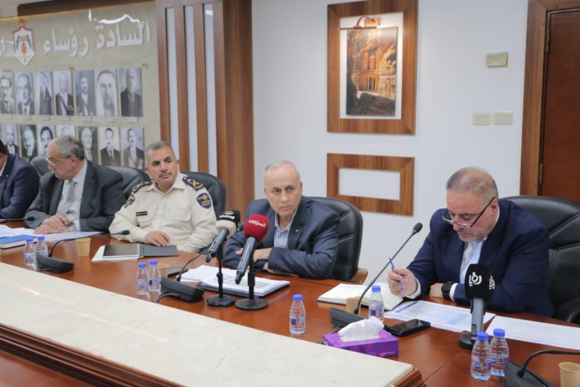 Officials sit at a conference table with microphones during a press briefing; Arabic posters and framed images hang on the wall behind them.