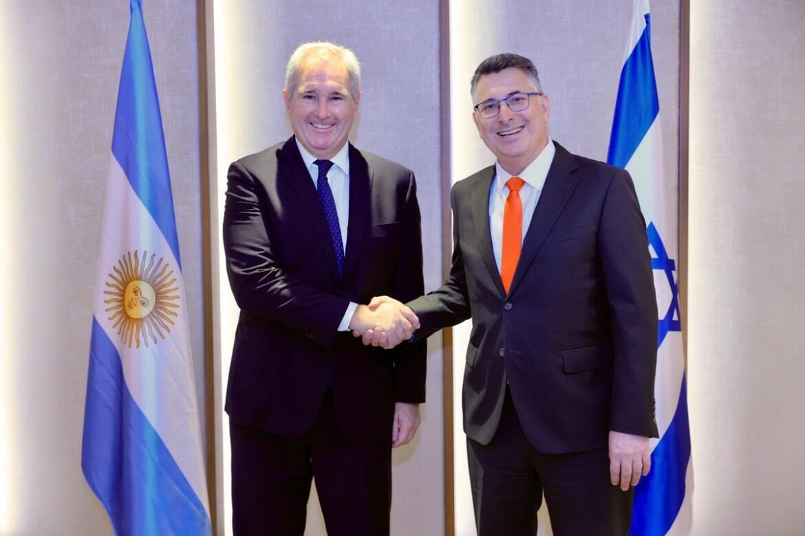 Two men in dark suits shake hands in front of Argentina and Israel flags.