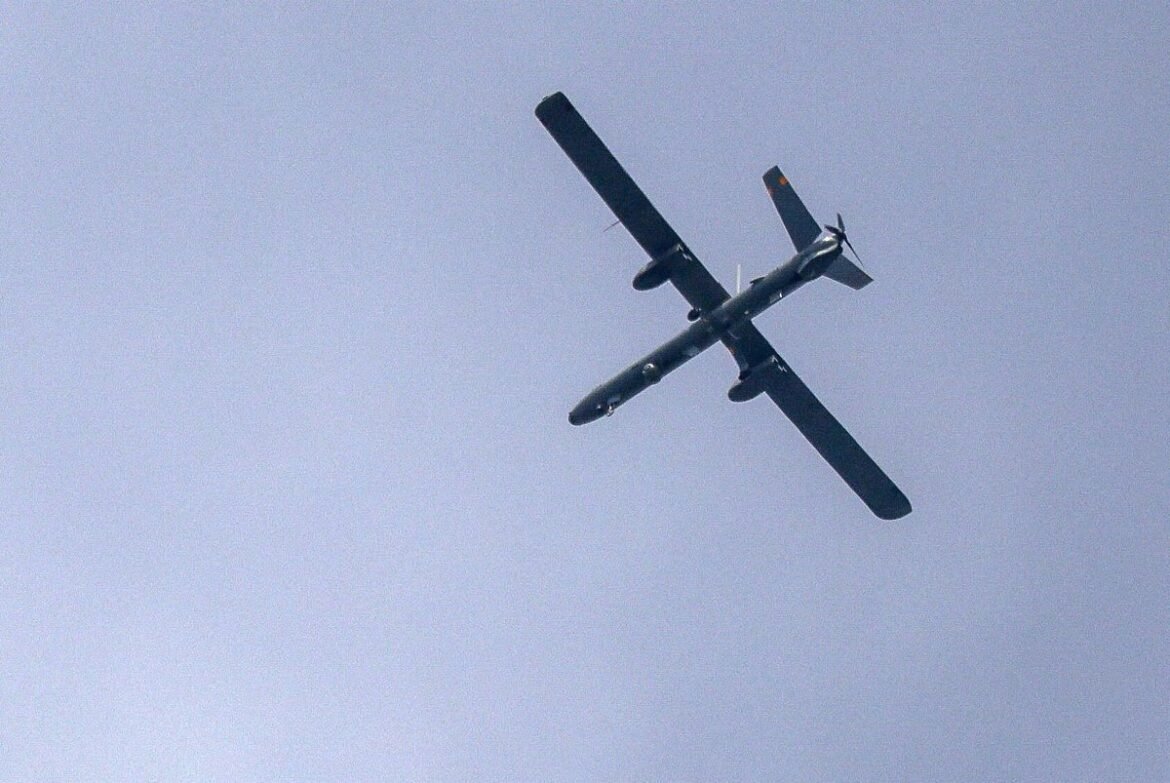 Twin-engine propeller airplane seen from below cruising in a clear blue sky, silhouetted against the light background, with landing gear retracted and wings spanning across the frame.