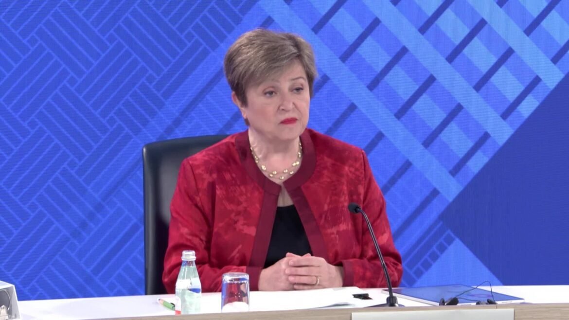 Woman in a red patterned blazer sits at a panel desk with a microphone and water bottle against a blue geometric backdrop.