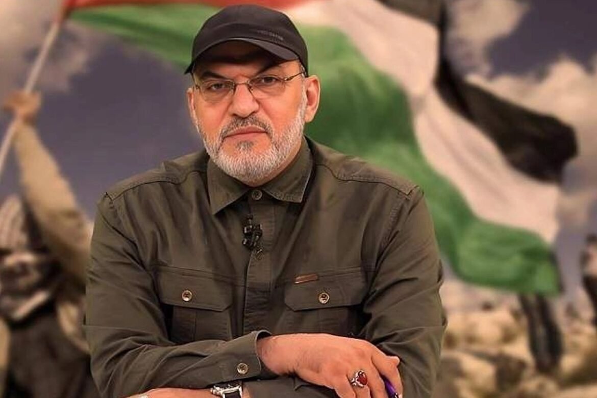 Man with a gray beard wearing a black cap and olive-green shirt, seated with a clip-on mic on his collar; a blurred flag backdrop behind him.