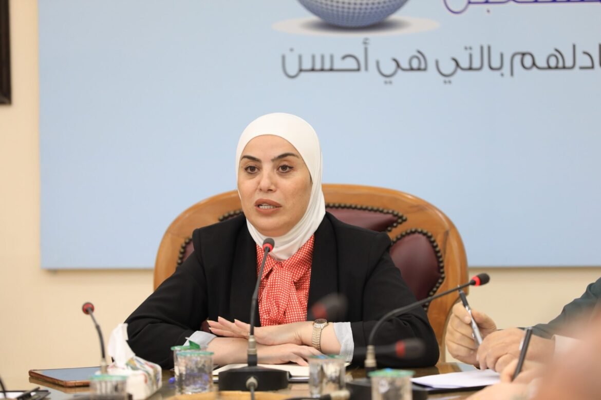 Woman in a white hijab speaking at a conference table with microphones and papers in front of her.