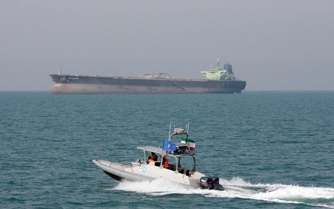 White motorboat with two people wearing orange life jackets speeds across turquoise water; a large cargo ship sits on the horizon behind.