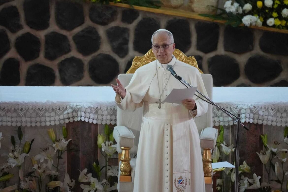 Pope in white robes speaks at a microphone while seated on a gilded throne, gesturing with one hand and holding papers.