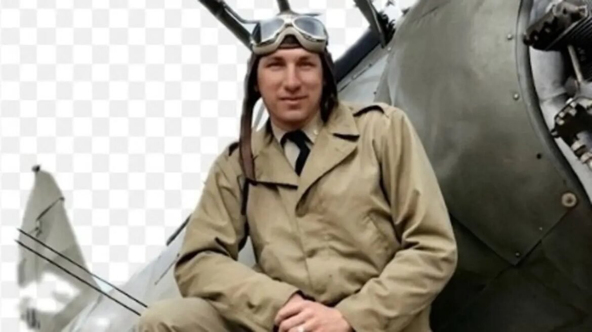 Man in vintage aviator outfit (khaki jumpsuit and goggles) sits beside a propeller airplane, posing for the photo.