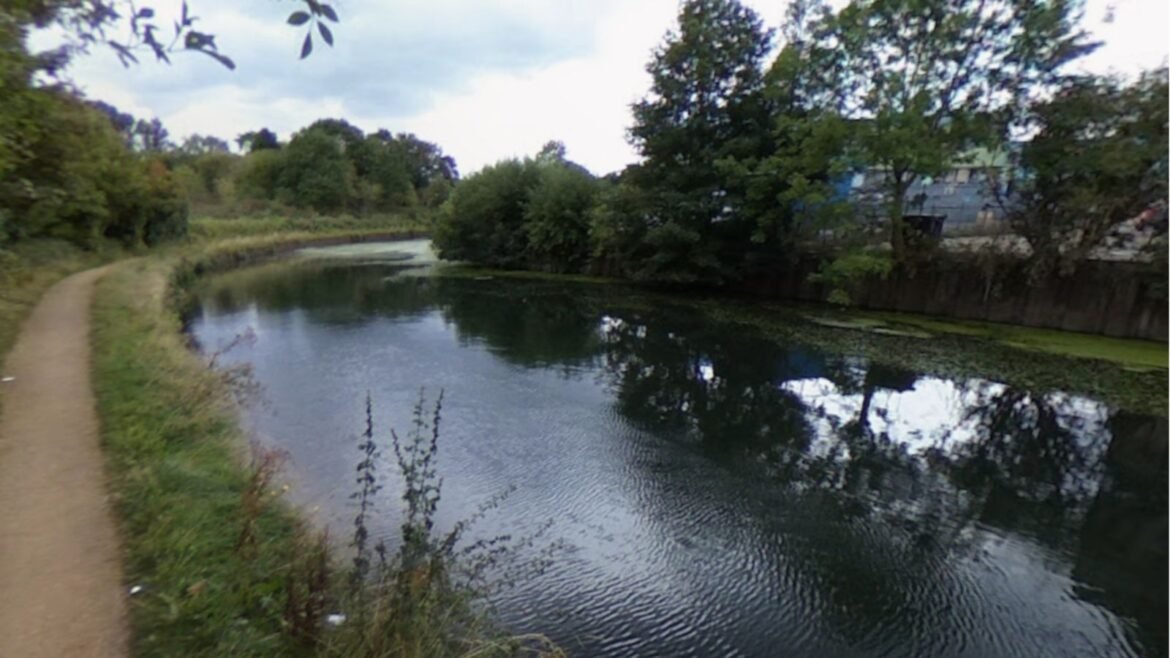 Dirt footpath runs along a calm canal, with trees and bushes on both sides and reflections on the water's surface.