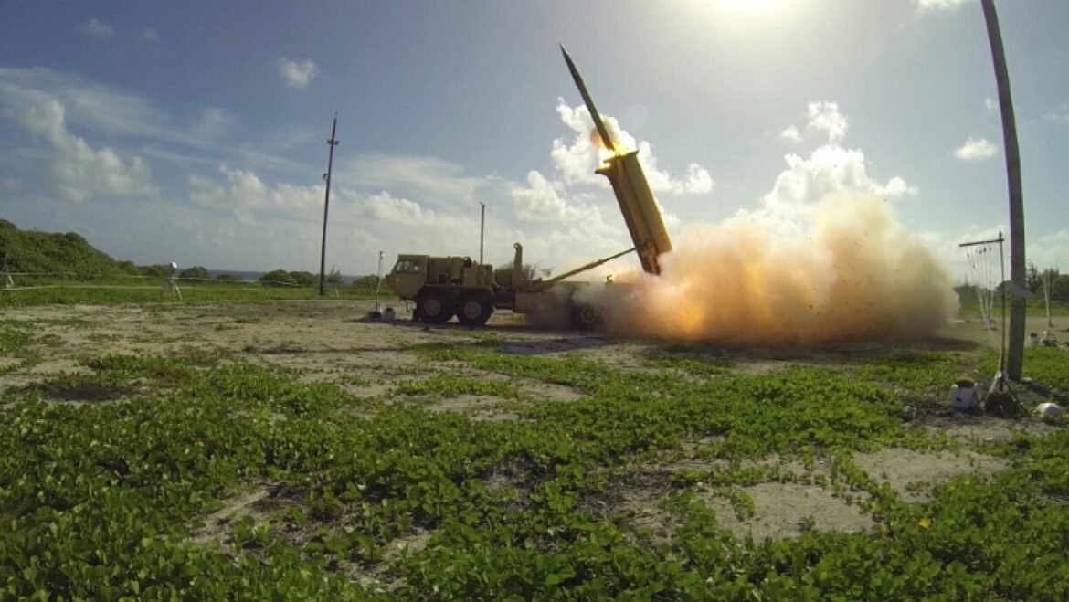 Mobile rocket launcher fires a missile on a sandy range; orange flame and a large dust cloud rise into a blue sky with scattered clouds.