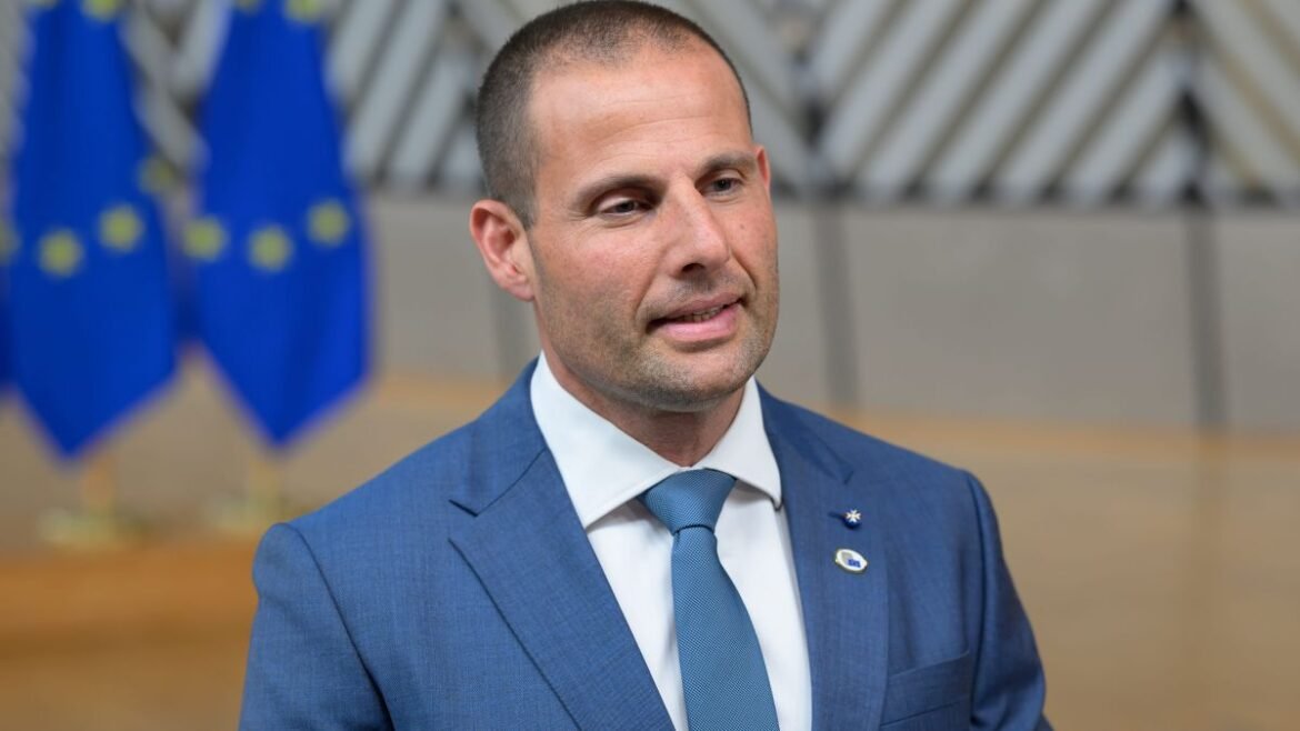 Man in a blue suit and tie speaks at a formal event, with European Union flags in the background.