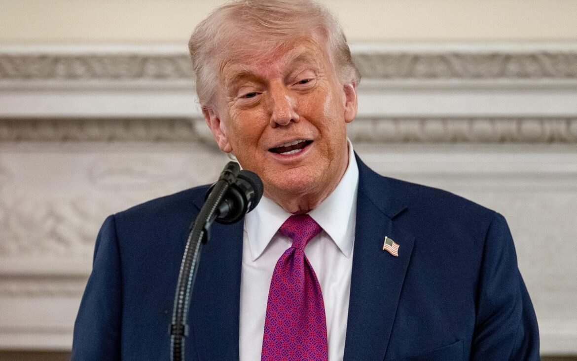 Man in a navy suit and magenta tie speaks into a microphone, wearing an American flag lapel pin at a formal event.