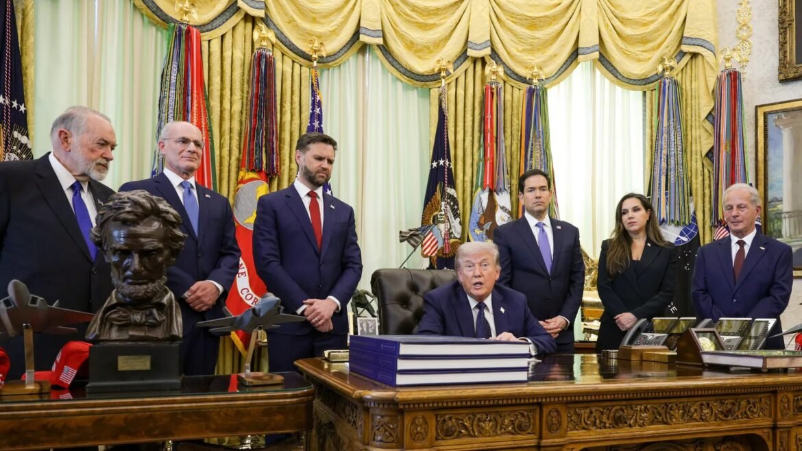 Donald Trump seated at a large desk in the ornate Oval Office, with several suited aides standing behind him.