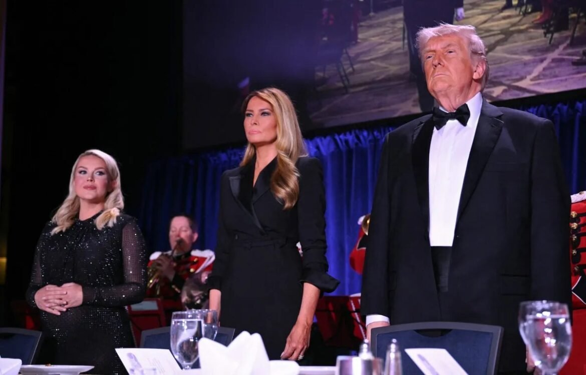 Three people in formal attire stand at a banquet table on a stage during a gala, with musicians in the background