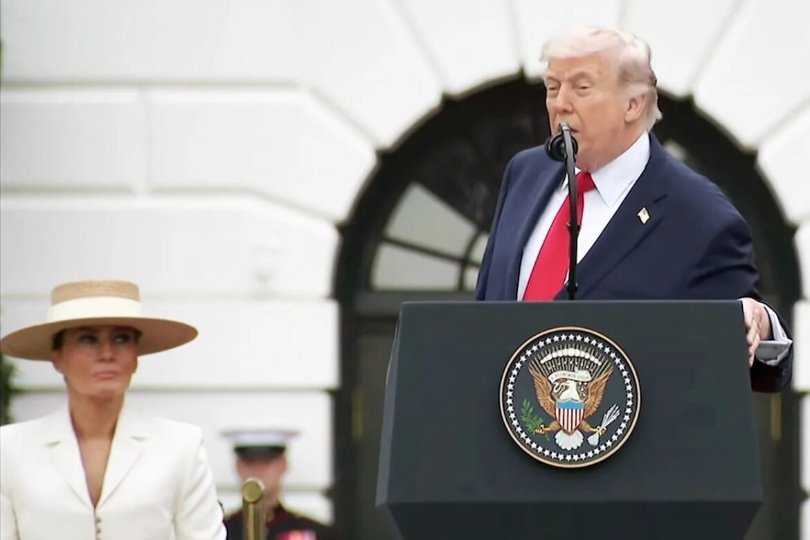 Man in a navy suit and red tie speaks at a presidential podium with the U.S. seal; a woman in white with a wide-brim hat stands nearby.