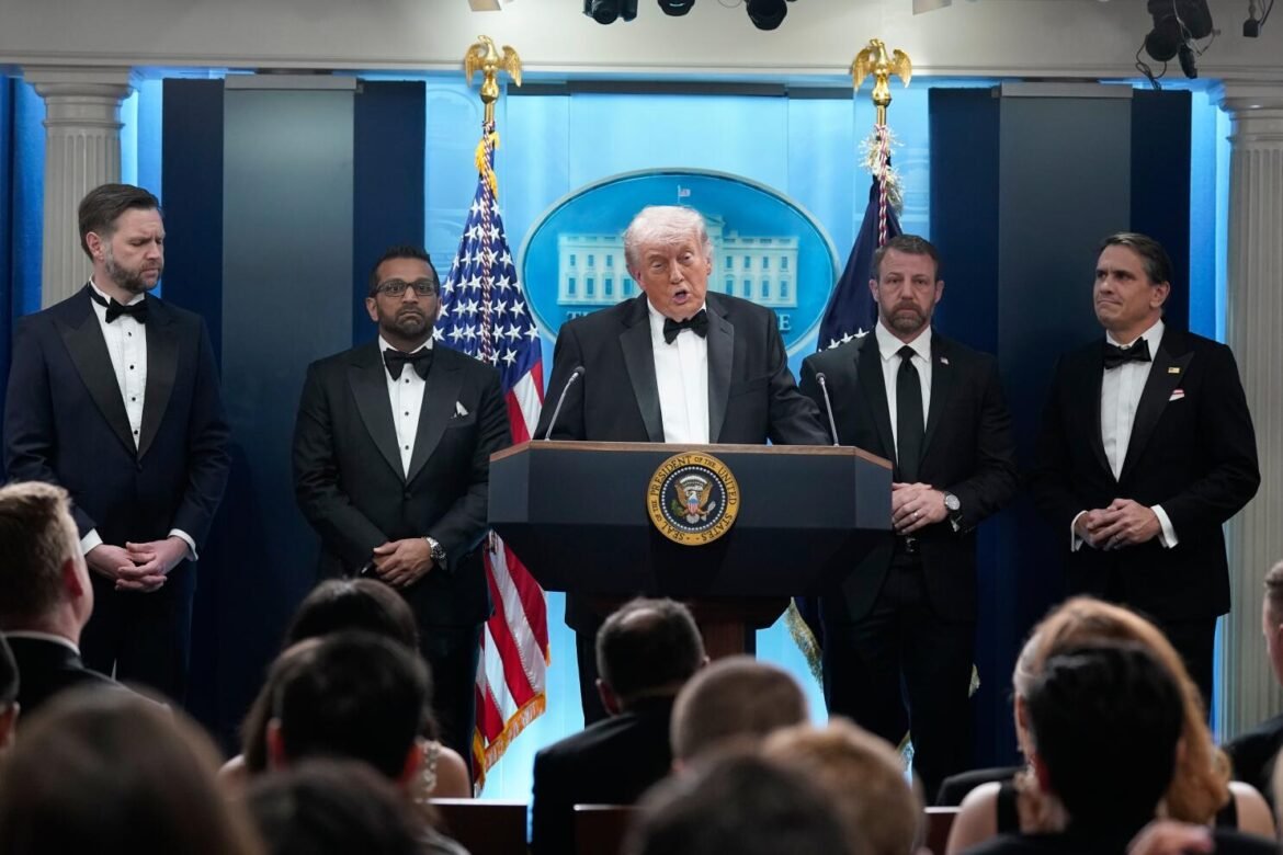 President at a podium delivering a speech, flanked by four men in tuxedos, with American flags in the background at a formal briefing.