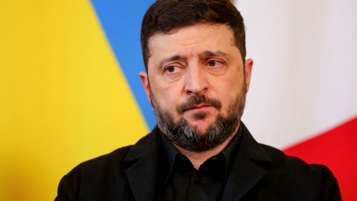 Close-up portrait of a man with a dark beard in a black suit, looking to the left, against a bright color-block background.