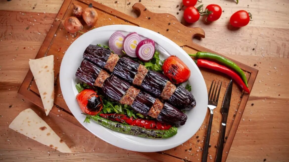 Plate of grilled eggplant rolls on a bed of greens with roasted tomatoes, onion slices, and peppers; cheese wedges on the side.