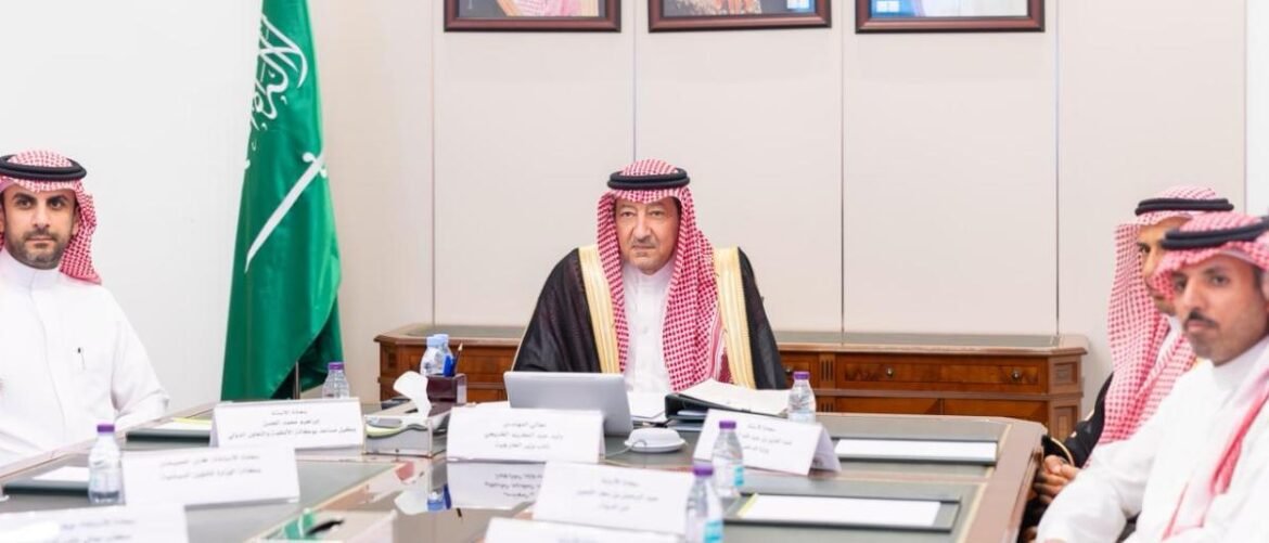 Central official in traditional Saudi attire sits at a conference table with laptops and documents; a green Saudi flag stands behind him in a formal meeting room.