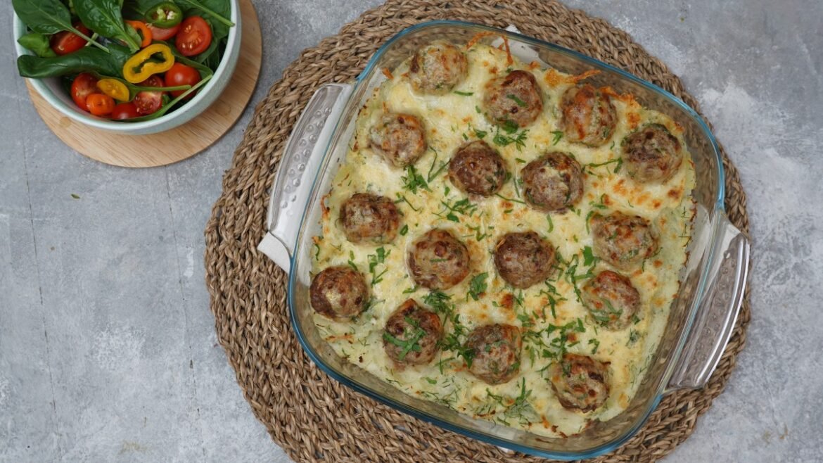 Baked meatballs in a creamy herb sauce in a glass dish on a woven placemat, with a side veggie salad nearby.