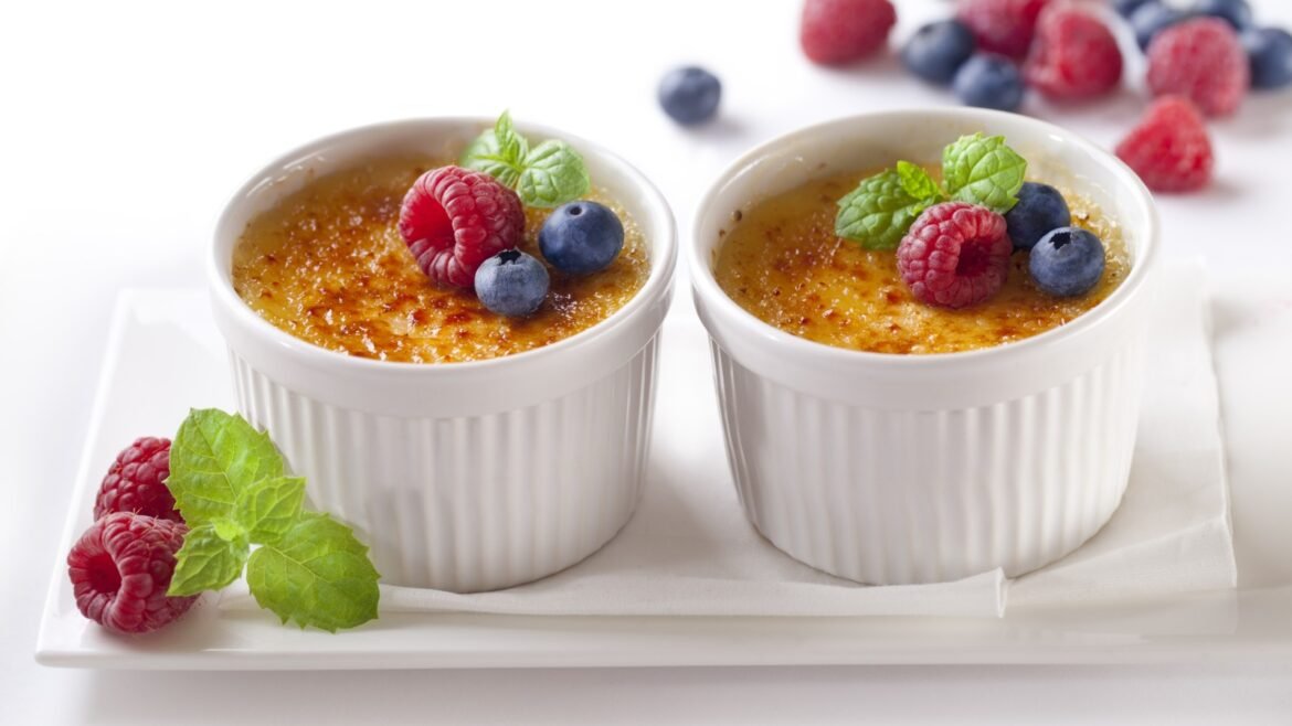 Two white ramekins of crème brûlée topped with caramelized sugar, garnished with raspberries, blueberries, and mint on a white plate.