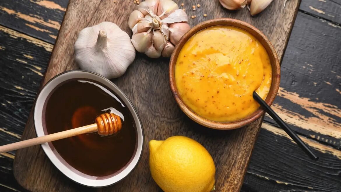 Rustic wooden board with garlic bulbs, a lemon, a honey pot and dipper, and a clay bowl of yellow mustard sauce.