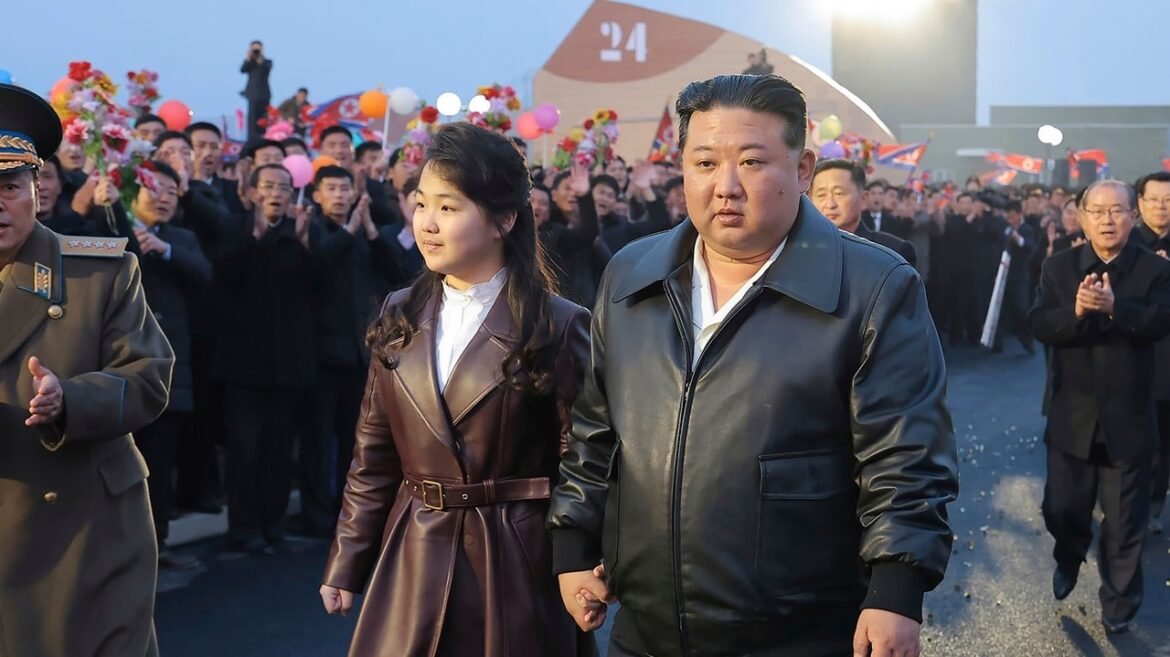 A man in a black leather jacket and a woman in a brown coat walk arm-in-arm through a cheering crowd with balloons and flags behind them.