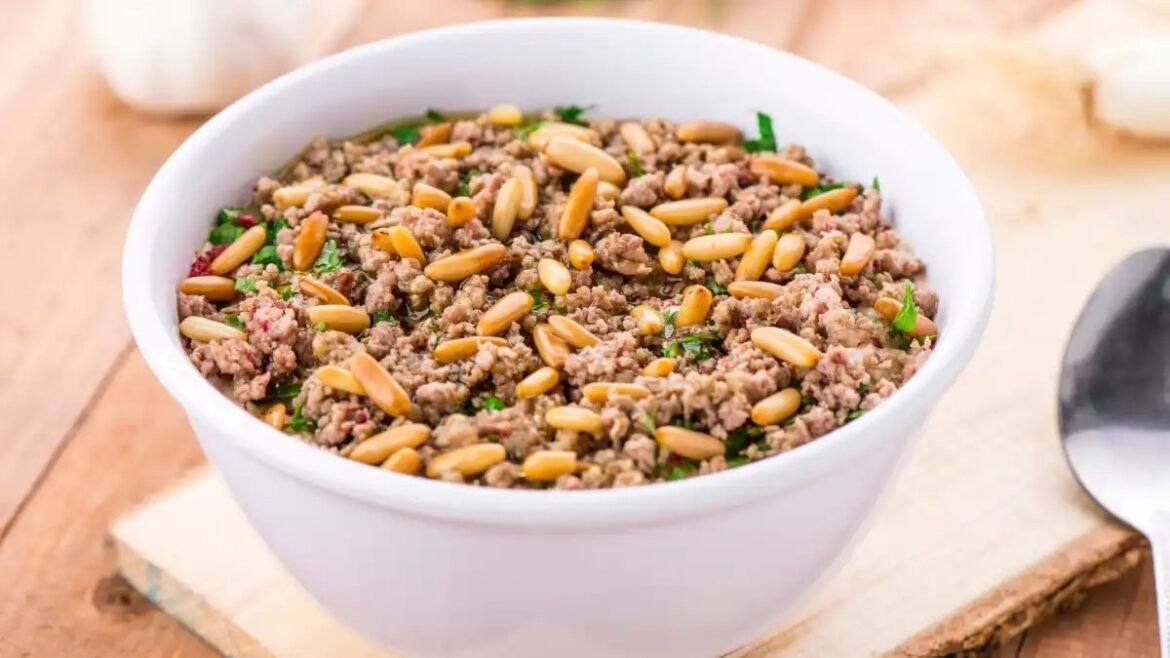 White bowl filled with cooked ground beef and pine nuts, garnished with parsley, on a wooden table.