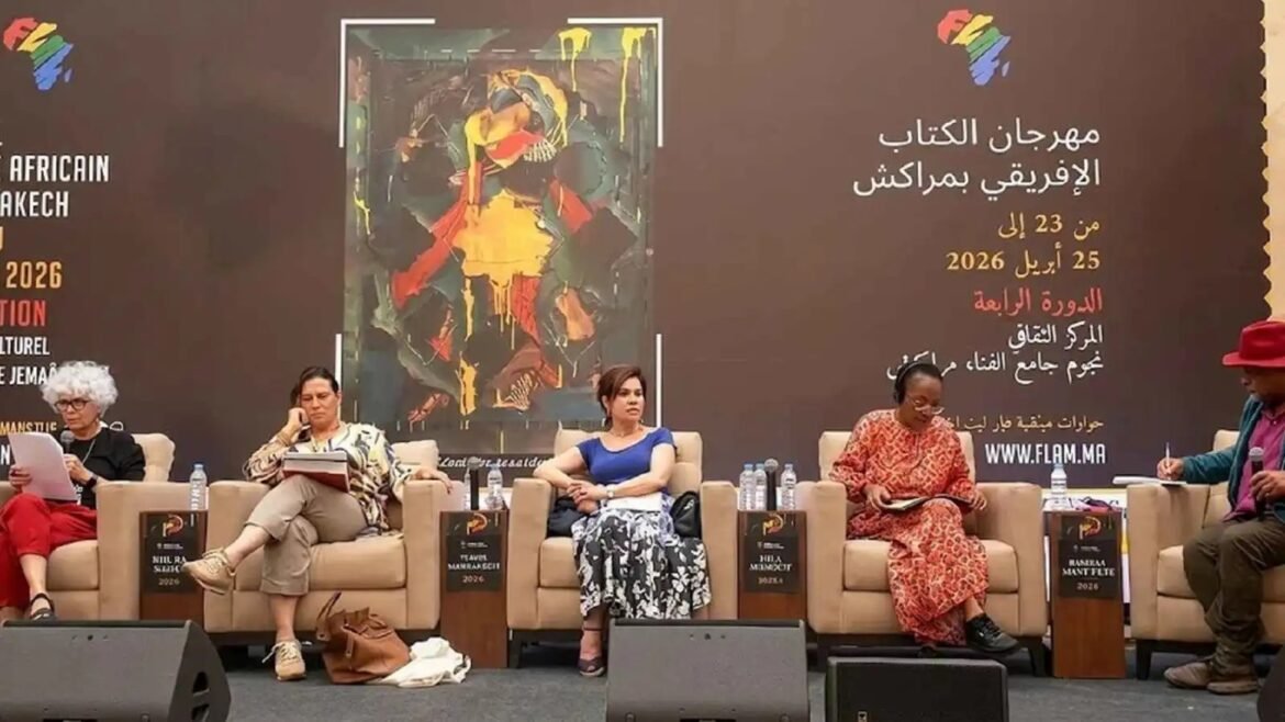 Panel discussion at a book festival stage with five speakers seated in beige chairs against a dark backdrop with Arabic text and artwork.