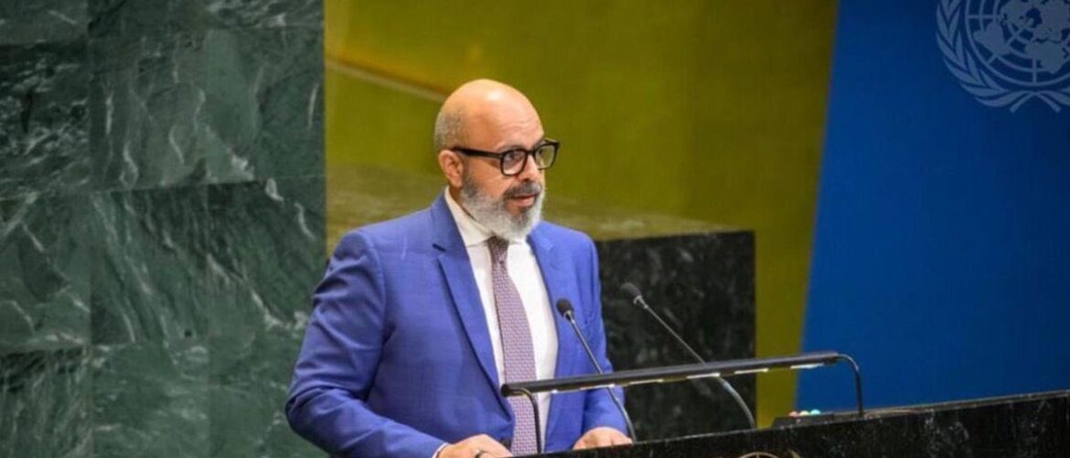 Speaker in a blue suit and glasses delivering a speech at a podium against a backdrop featuring the UN emblem and dark marble panels.