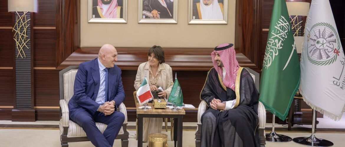Three people sit and chat in a formal meeting room; a man in a blue suit, a woman in beige, and a man in traditional Saudi attire, with Kuwaiti and Saudi flags on the table.