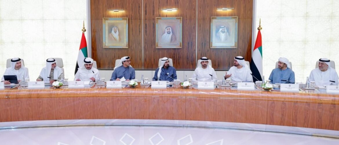 Dignitaries in traditional Gulf attire seated at a curved wooden conference table with UAE flags and framed portraits behind them.