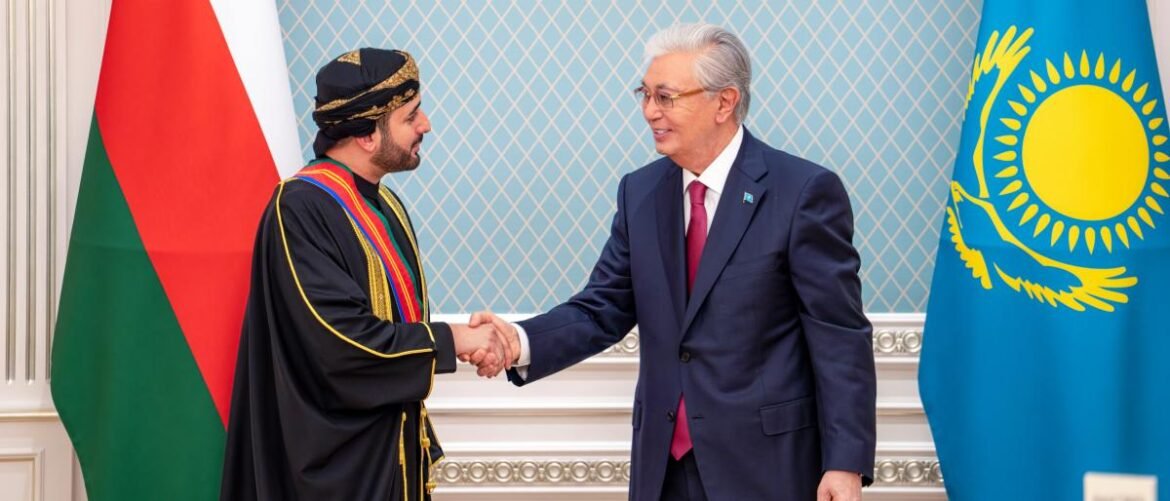 Two men shake hands in a formal room: one in traditional Omani attire, the other in a dark suit, with the flags of Oman and Kazakhstan in the background.