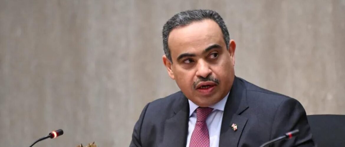 Man in a dark suit and pink tie speaking at a panel, with microphones in front of him against a plain background.