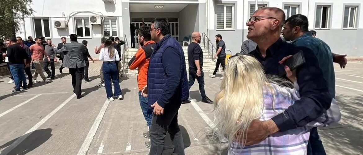 Group of adults standing and walking in a parking lot outside a white building; a man hugs a blonde woman in the foreground.