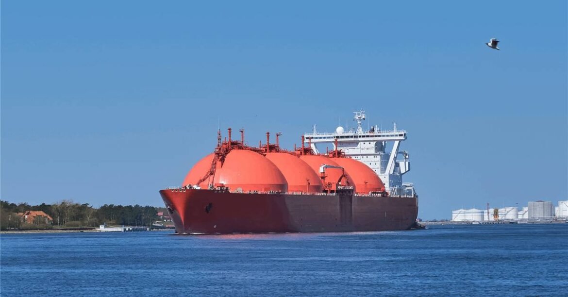 Red LNG carrier with three spherical tanks sailing on blue water toward a port.