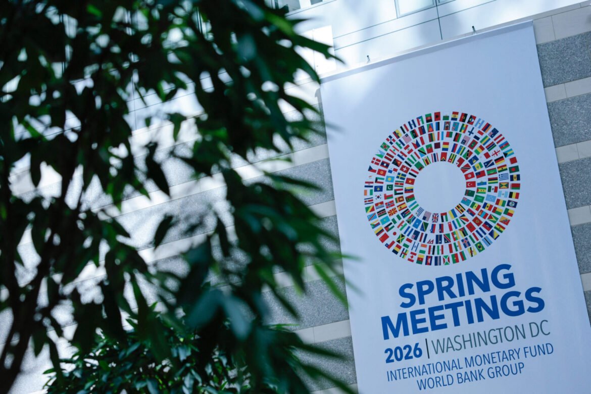 Banner announcing Spring Meetings 2026 in Washington DC, featuring a circular mosaic of national flags and IMF/World Bank text (partially obscured by leaves).