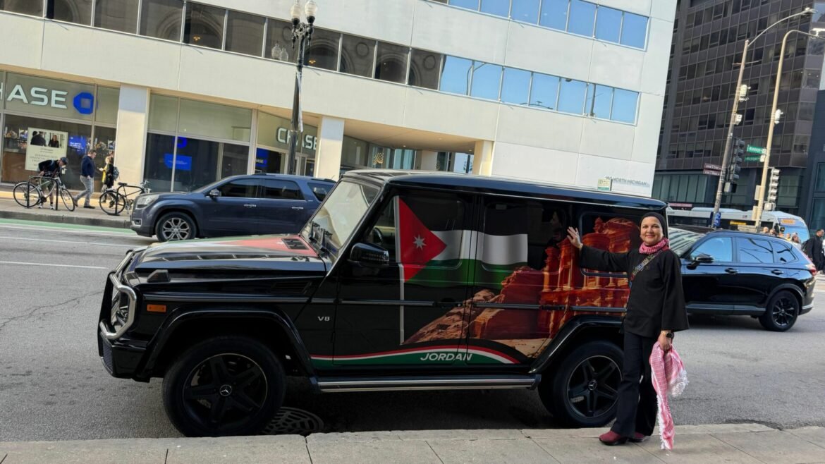 Woman in a headscarf waves beside a black Mercedes SUV wrapped in the Jordan flag on a city street.