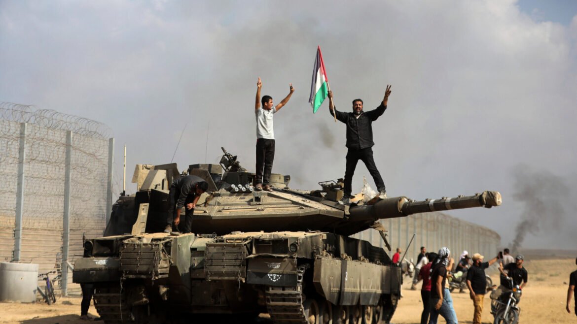 Two men stand on a tank in a desert area; one waves a Palestinian flag while others gather nearby near a barbed-wire fence.
