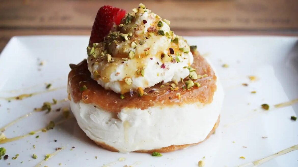 Round dessert on a biscuit base with thick white cream, topped with whipped cream, crushed pistachios and a strawberry garnish