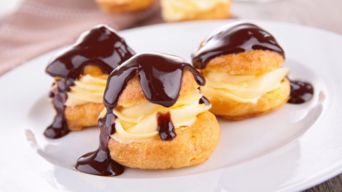 Three cream-filled choux pastries topped with glossy chocolate sauce on a white plate, a dessert trio.